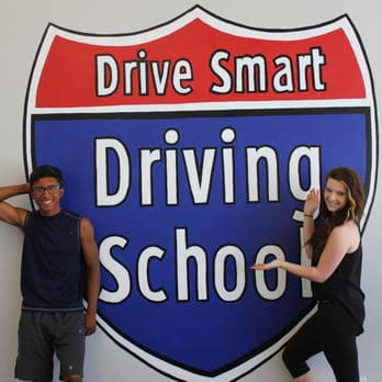 Two people beside the Drive Smart Driving School shield at the school